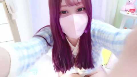 Media: A video of a young woman with long, straight purple hair, wearing a white face mask, a blue and white plaid robe, and a white tank top, taking a selfie in a bright, soft-focused bathroom.