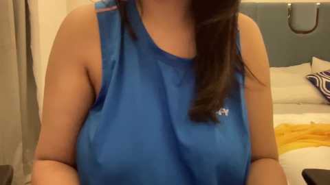 Media: Video of a woman in a blue sports tank top, with medium build and medium-sized breasts, sitting on a bed with white sheets and a grey headboard. Her long, straight, dark hair falls over her shoulder.