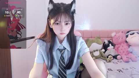 Media: A video of a young East Asian woman with long black hair and cat ears, wearing a blue shirt and striped tie, sitting on a bed with stuffed animals in a pink room.