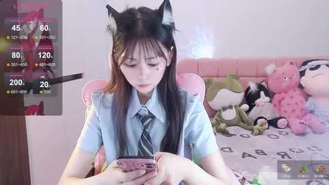 Media: Video of a young Asian woman with long black hair, wearing cat ears, blue shirt, and striped tie, playing with a pink smartphone, surrounded by plush toys in a pink room.