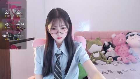 Media: A video of an Asian woman with long black hair and glasses, wearing a light blue shirt and striped tie, sitting in a pink gaming chair, surrounded by stuffed animals in a cozy bedroom.