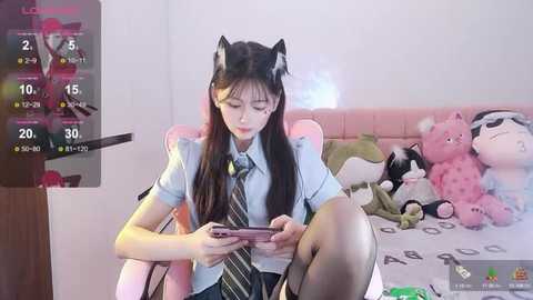 Media: Video of an East Asian woman in a cat costume, sitting on a pink chair, playing with a smartphone. Background includes a bed with stuffed toys and a monitor displaying a video game.