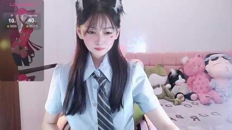 Media: Video of a young East Asian girl with long black hair, wearing a light blue school uniform, standing in a bedroom with stuffed animals and a pink wall.