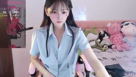 Media: Video of an East Asian woman with long black hair, wearing a light blue button-up shirt, sitting on a pink chair in a bedroom with plush toys, a bed, and a wooden wardrobe.