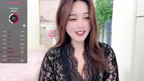 Media: Video of a young East Asian woman with long brown hair, wearing a black lace robe, smiling in a brightly lit room with a potted plant and wall art in the background.