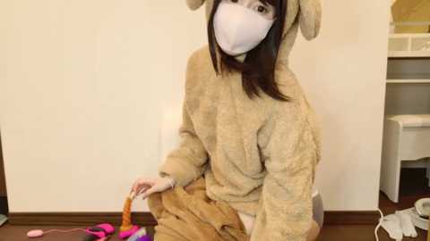 Media: A young Asian woman with long black hair and a white mask, dressed in a plush rabbit costume, sits on the floor, holding a pink wand, in a sparsely furnished room with a white wall and wooden floor.