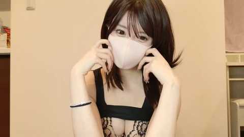 Media: Video of a young Asian woman with shoulder-length black hair, wearing a black lace bra, holding a pink face mask. Background shows a beige wall and a white shelf with items.