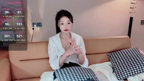 Media: Video of an East Asian woman with fair skin, long black hair, and red lipstick, wearing a white blouse, sitting on a beige couch. Background features a flat-screen TV showing weather forecasts.