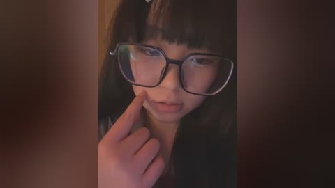 Media: A video of a young Asian woman with straight black hair and large rectangular glasses, lightly touching her cheek, set against a warm, dimly-lit background.