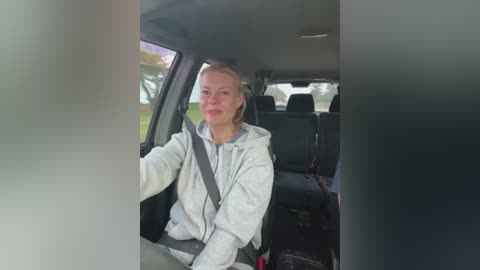 Media: Video of a blonde woman in a light gray hoodie, driving a black car, with a black seatbelt on, inside a dimly lit vehicle.