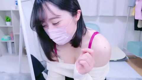 Media: Video of an East Asian woman with straight black hair, wearing a white off-shoulder top, pink bra, and face mask, sitting in a modern, clean room with white walls and minimal decor.