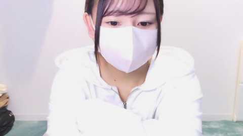 Media: A video of an Asian woman with short dark hair, wearing a white face mask, white hoodie, and light blue pants, sitting on a green carpet in a plain white room.