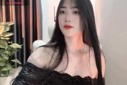 Media: Video of an East Asian woman with long black hair, pale skin, red lipstick, and a black lace off-shoulder top, indoors with a potted plant and vertical blinds in the background.