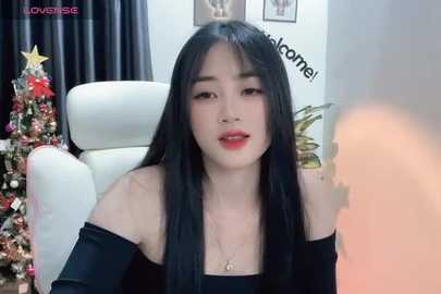 Media: A video of an Asian woman with long black hair, wearing a black off-shoulder top, seated in a white chair in a cozy room with a decorated Christmas tree and framed artwork on the wall.