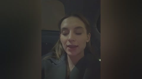 Media: A dimly-lit video of a woman with light skin, brown hair, and a gray blazer, eyes closed, smiling, possibly in a car.