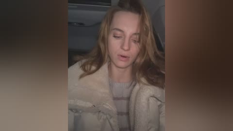 Media: Video of a young woman with light skin, long blonde hair, and closed eyes, wearing a beige fur coat and a striped sweater. She appears to be sitting in a car.