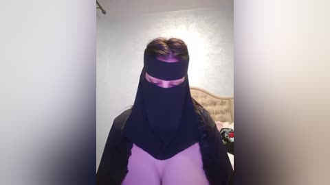 Media: A video of a person wearing a black niqab, obscuring their face, standing in a dimly lit room with a beige headboard and a pinkish-purple wall.