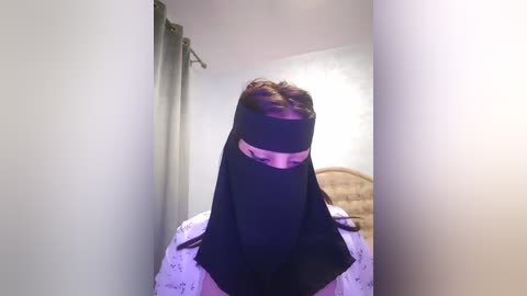 Media: Video of a woman wearing a black niqab with a pink stripe across the eyes, sitting on a bed with a white floral sheet, surrounded by white walls and green curtains in a dimly lit room.