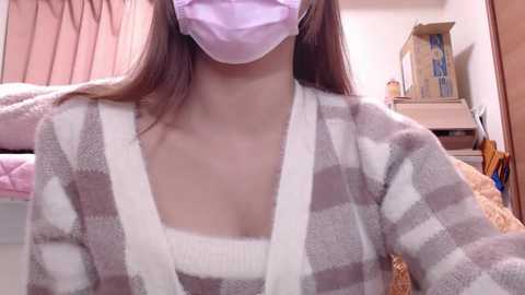 Media: Video of an Asian woman wearing a purple face mask and a light purple top, with a beige and white checkered cardigan, in a cozy, cluttered bedroom with pink curtains and a wooden cabinet.