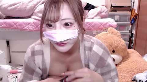 Media: A video of an Asian woman with light skin, wearing a face mask, striped pajama top, and holding a brush. She sits on a pink bed with a teddy bear, in a cluttered room.