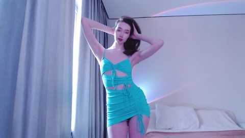 Media: Video of a slim, fair-skinned woman with long dark hair, wearing a turquoise, ruched mini-dress, posing provocatively in a modern bedroom with white walls and a bed.