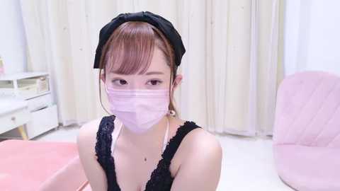 Media: Video of an Asian woman with light skin, wearing a black headband, black lace top, and pink face mask, sitting in a pink chair, in a minimalistic room with white walls and curtains.