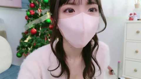 Media: A video of an Asian woman with long, wavy black hair and pale skin, wearing a white face mask, standing in front of a decorated Christmas tree and a white dresser.