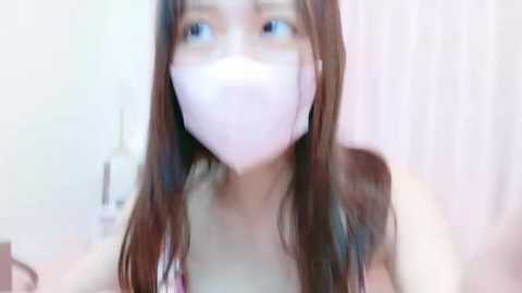 Media: Video of an Asian woman with long brown hair, wearing a white face mask, looking towards the camera. The background is a softly blurred, light-colored room with vertical blinds.