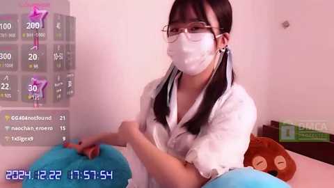 Media: Video of an East Asian woman in a white lab coat and mask, sitting on a bed with a blue pillow, wearing glasses. Background shows a digital clock and social media icons.