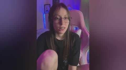 Media: Video of a young woman with glasses and straight brown hair, wearing a black t-shirt, sitting on a pink gaming chair in a dimly lit room with blue LED lights.