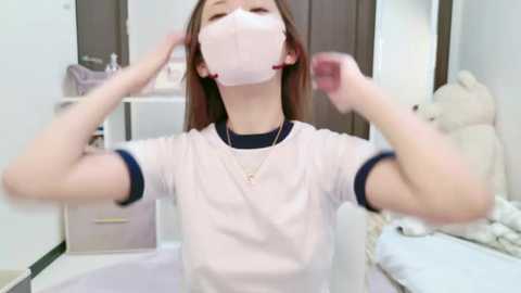 Media: Video of an Asian woman with long brown hair, wearing a white T-shirt and pink face mask, posing with hands on head in a bright, clean bedroom with white walls, a bed, and stuffed animals.