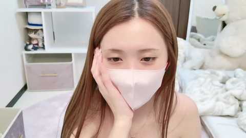 Media: A video of an Asian woman with long, straight brown hair, wearing a white face mask, sitting on a bed in a bedroom with white furniture and stuffed animals.