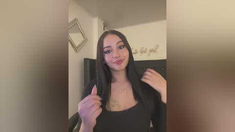 Media: Video of a young woman with long black hair, fair skin, and a slender physique, wearing a black top. She gives a thumbs-up sign in a modestly decorated room with beige walls and a framed quote.