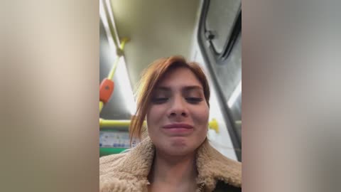 Media: A video of a Caucasian woman with fair skin and red hair, wearing a beige fur-trimmed jacket, smiling and looking out the window of a bus. The background shows a blurred interior with yellow and orange seats.