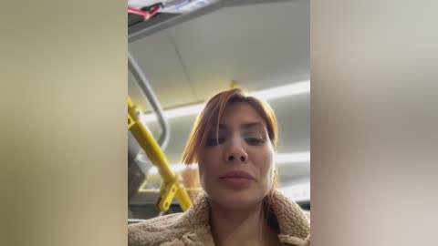 Media: Video of a woman with light brown hair, wearing a beige coat, sitting on a yellow seat in a dimly lit train compartment.