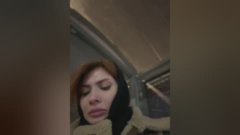 Video of a young woman with fair skin and shoulder-length auburn hair, wearing a black hooded jacket, looking downcast, seated in a car. The image is dimly lit with a muted color palette.