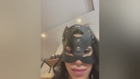 Media: Video of a woman with light skin and long dark hair wearing a black Batman mask, in a modern, minimalist room with white walls and recessed lights.