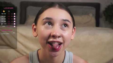 Media: Video of a young woman with fair skin, dark hair, and a nose ring, sticking out her tongue in a playful manner. She wears a gray tank top. Background shows a bed with beige bedding and a green plant.