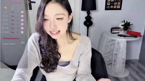 Media: Video of an Asian woman with long black hair, wearing a grey cardigan and grey bra, smiling playfully in a modern, minimalist room with white walls, a round table, and a black lamp.