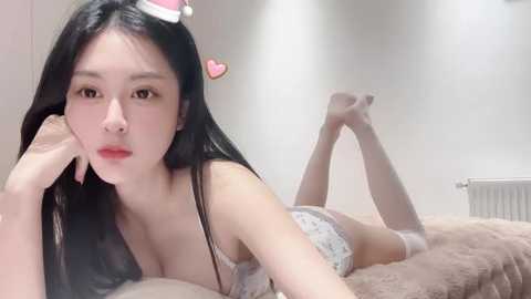 Media: Video of an East Asian woman with long black hair and fair skin, lying on her stomach in lingerie, in a bedroom with a beige bedspread and white walls.