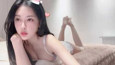 Media: A video of an Asian woman with long black hair, lying on a beige bed in a bedroom, wearing a lacy white bra and panties, with a pink bow in her hair.