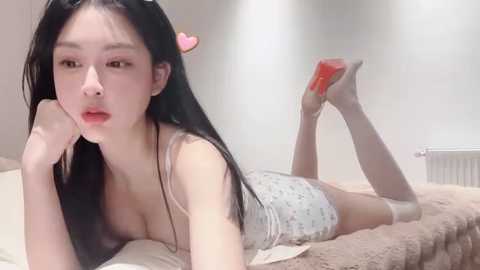 Media: Video of an Asian woman with long black hair, fair skin, and a slender physique, lying on a bed in a white room, wearing a white camisole and shorts, holding a red phone, and playfully wiggling her legs.