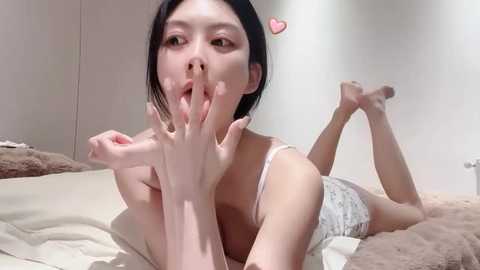 Media: Video of an East Asian woman with straight black hair, lying on a bed in a white room, wearing a white lace bra and panties. She is playfully covering her mouth and making a heart gesture with her fingers.