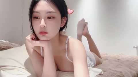 Media: Video of a young Asian woman with fair skin, black hair, and pink lipstick, lying on a beige bed in a minimalist room, wearing a white lace bra and panties.