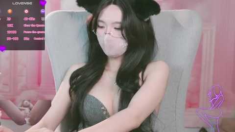 Media: A video of an Asian woman with long black hair, wearing a grey lace dress and black cat ears, sitting on a grey chair with a pink mask. Background shows a soft pink room with heart icons.