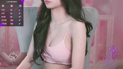 Media: Video of an East Asian woman with long, wavy black hair, wearing a pink ribbed crop top, sitting indoors with pink walls and a chair.