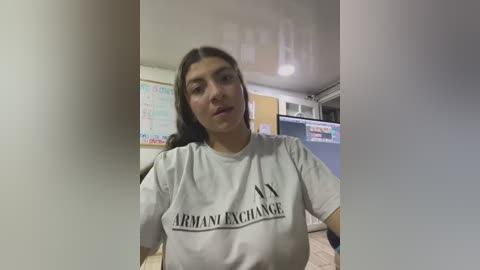 Media: Video of a young woman with dark hair, wearing a white T-shirt with \"ARMY\" and \"ARMY EXCHANGE\" printed in black letters, sitting in a classroom with posters on the walls.