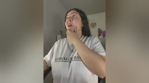 Media: A video shows a Caucasian woman with long dark hair, wearing a white T-shirt, thinking with her hand on her chin, in a dimly lit room with a decorative heart on the wall.