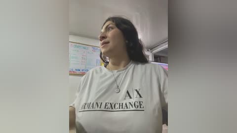 Media: Video of a woman with long dark hair, wearing a white t-shirt with \"AX\" and \"PANY EXCHANGE\" printed on it, standing indoors.