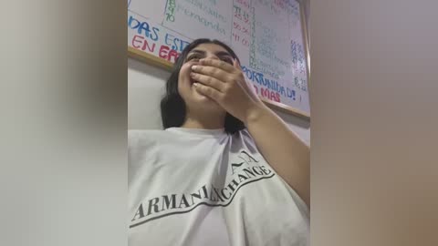 Media: Video of a young woman with medium brown skin, wearing a white t-shirt with \"ARMANIA\" in black, covering her mouth with her hand. Background features a chalkboard menu in various languages.